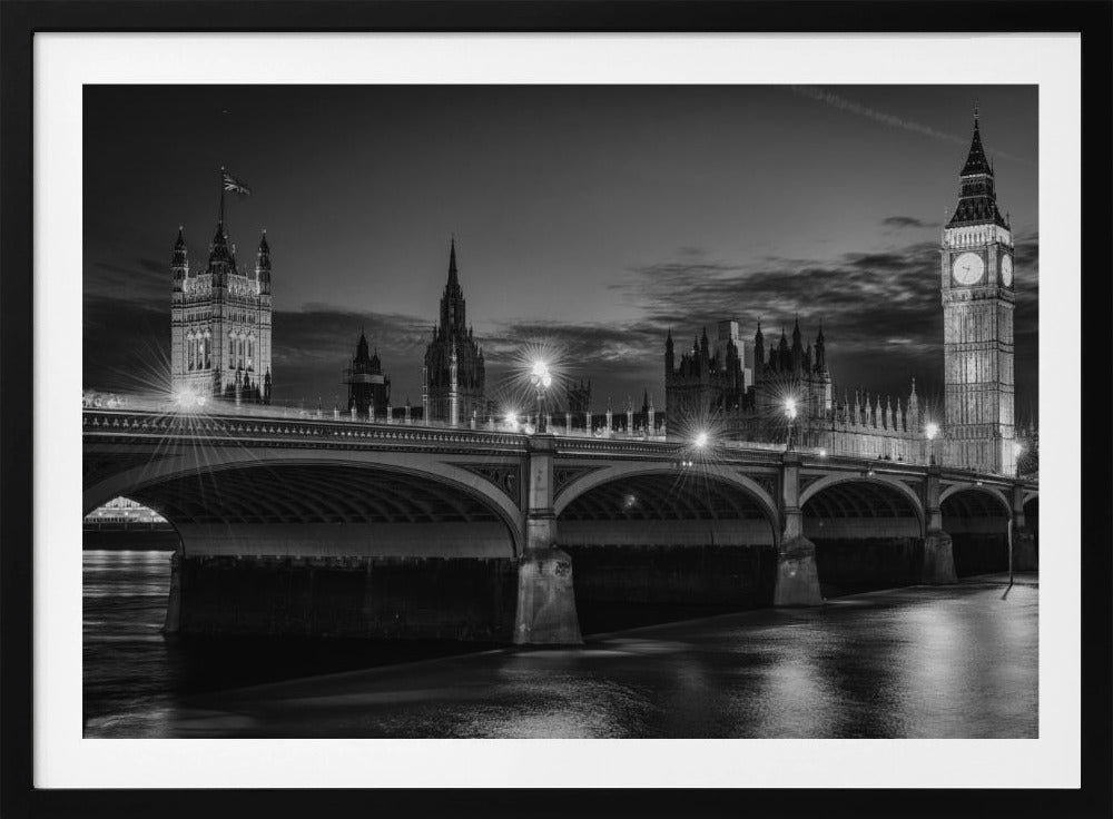 A framed, black and white nighttime photograph of Westminster Bridge in London. The iconic Houses of Parliament and the glowing Big Ben clock tower stand prominently in the background against a dark, cloudy sky. The streetlights on the bridge create starburst effects, and their reflections shimmer on the surface of the River Thames in the foreground. Poster