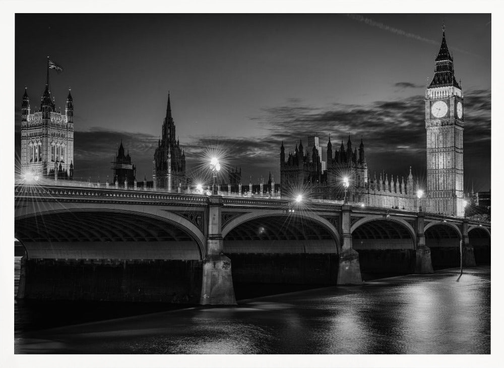 A framed, black and white nighttime photograph of Westminster Bridge in London. The iconic Houses of Parliament and the glowing Big Ben clock tower stand prominently in the background against a dark, cloudy sky. The streetlights on the bridge create starburst effects, and their reflections shimmer on the surface of the River Thames in the foreground. Poster