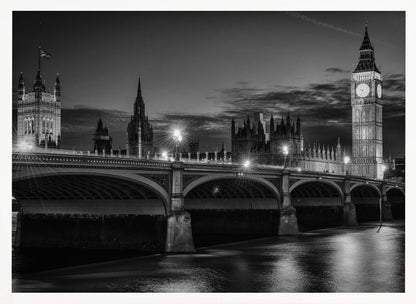 A framed, black and white nighttime photograph of Westminster Bridge in London. The iconic Houses of Parliament and the glowing Big Ben clock tower stand prominently in the background against a dark, cloudy sky. The streetlights on the bridge create starburst effects, and their reflections shimmer on the surface of the River Thames in the foreground. Poster