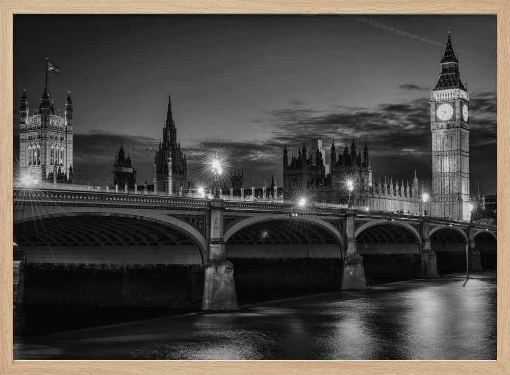 A framed, black and white nighttime photograph of Westminster Bridge in London. The iconic Houses of Parliament and the glowing Big Ben clock tower stand prominently in the background against a dark, cloudy sky. The streetlights on the bridge create starburst effects, and their reflections shimmer on the surface of the River Thames in the foreground. Poster
