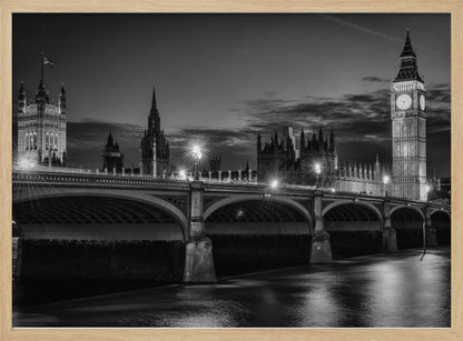 A framed, black and white nighttime photograph of Westminster Bridge in London. The iconic Houses of Parliament and the glowing Big Ben clock tower stand prominently in the background against a dark, cloudy sky. The streetlights on the bridge create starburst effects, and their reflections shimmer on the surface of the River Thames in the foreground. Poster