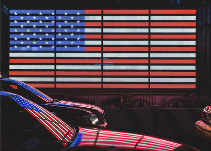 A large illuminated American flag on a billboard at night, with its red, white, and blue lights reflecting on the glossy hoods of two cars in the foreground. Wall Art