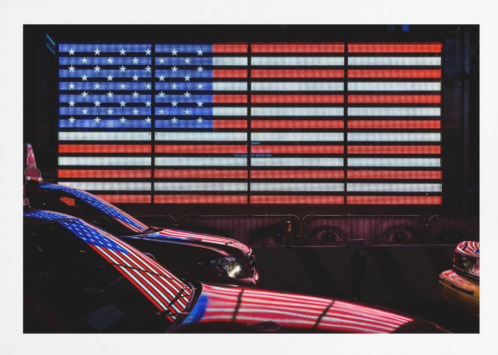 A large illuminated American flag on a billboard at night, with its red, white, and blue lights reflecting on the glossy hoods of two cars in the foreground. Wall Art