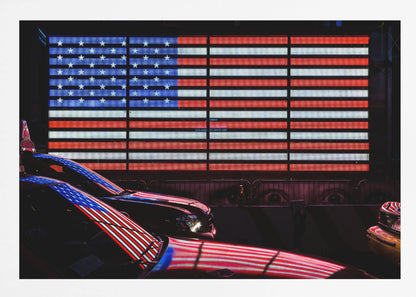 A large illuminated American flag on a billboard at night, with its red, white, and blue lights reflecting on the glossy hoods of two cars in the foreground. Wall Art