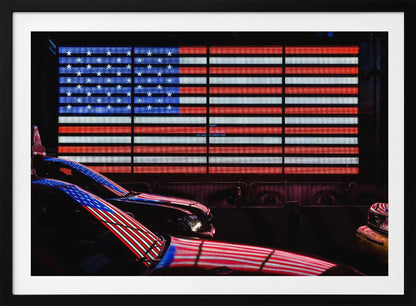 A large illuminated American flag on a billboard at night, with its red, white, and blue lights reflecting on the glossy hoods of two cars in the foreground. Wall Art