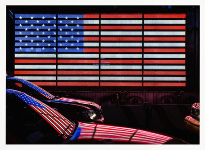 A large illuminated American flag on a billboard at night, with its red, white, and blue lights reflecting on the glossy hoods of two cars in the foreground. Wall Art
