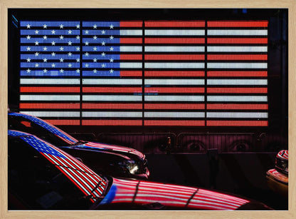 A large illuminated American flag on a billboard at night, with its red, white, and blue lights reflecting on the glossy hoods of two cars in the foreground. Wall Art