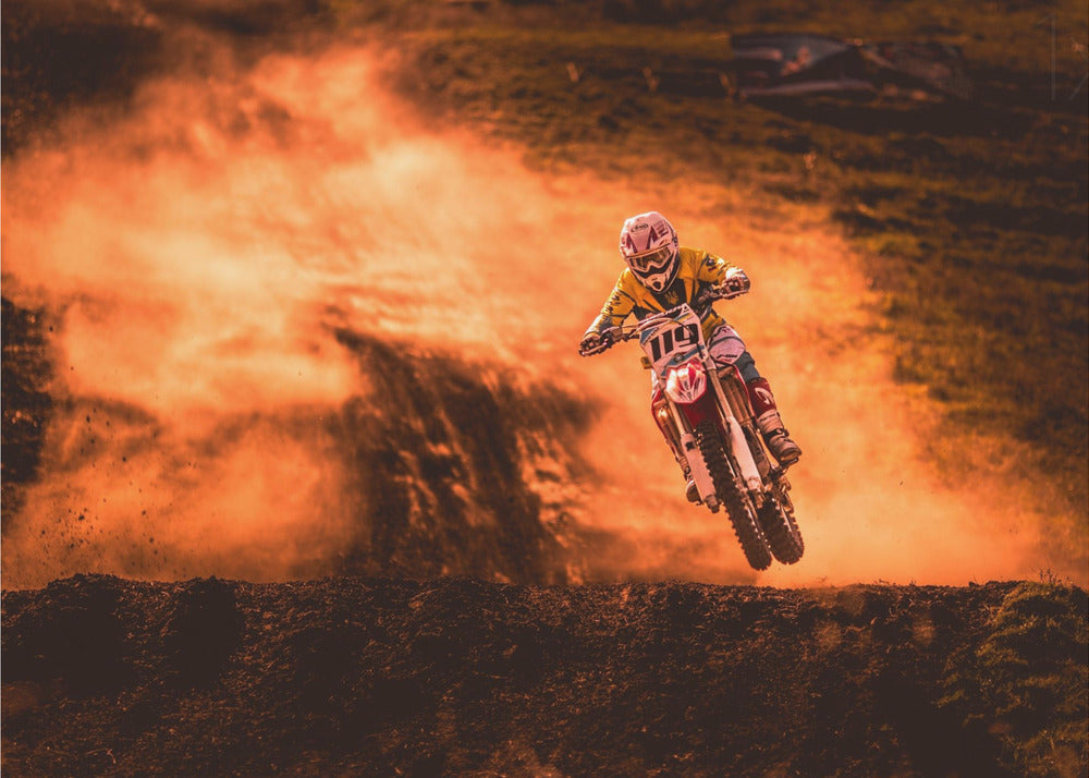 An action-packed photo of a motocross rider in mid-air, jumping their dirt bike over a mound of earth. A large cloud of orange dust billows behind them, creating a dramatic and fiery background. Wall Art