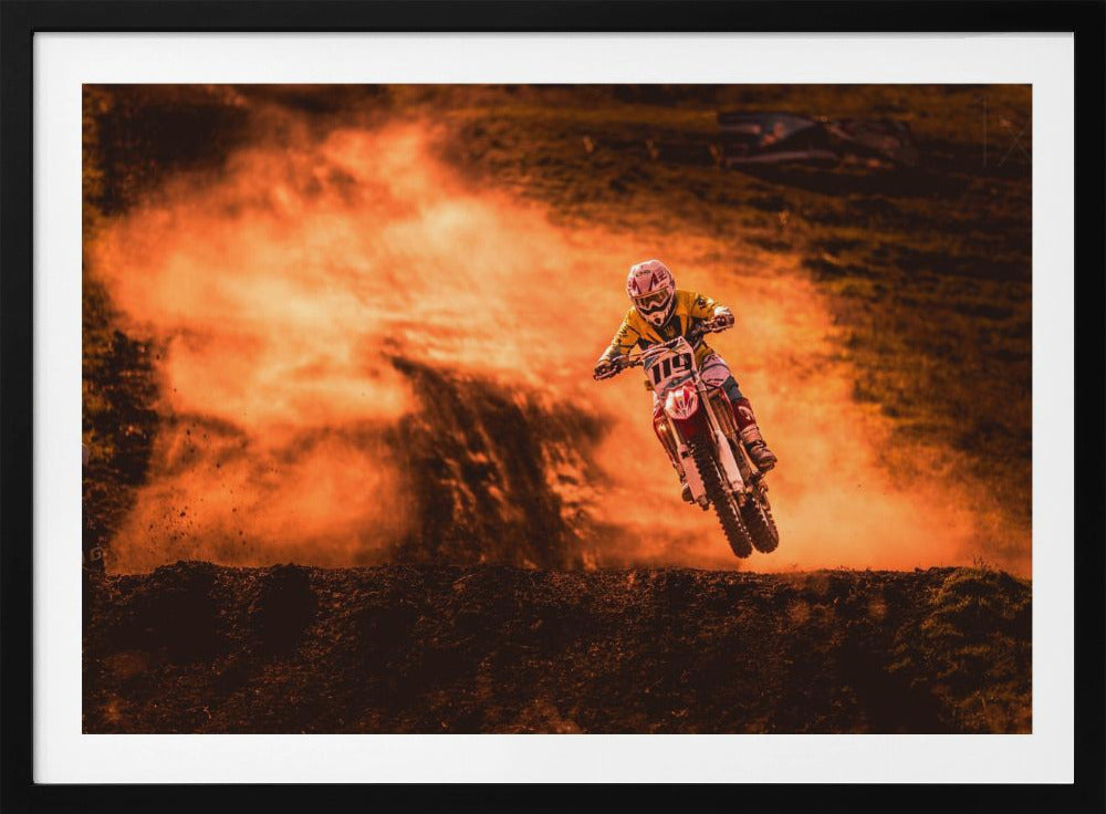 An action-packed photo of a motocross rider in mid-air, jumping their dirt bike over a mound of earth. A large cloud of orange dust billows behind them, creating a dramatic and fiery background. Wall Art