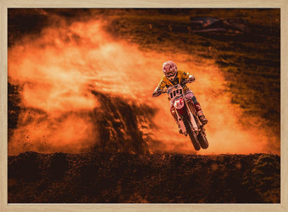 An action-packed photo of a motocross rider in mid-air, jumping their dirt bike over a mound of earth. A large cloud of orange dust billows behind them, creating a dramatic and fiery background. Wall Art