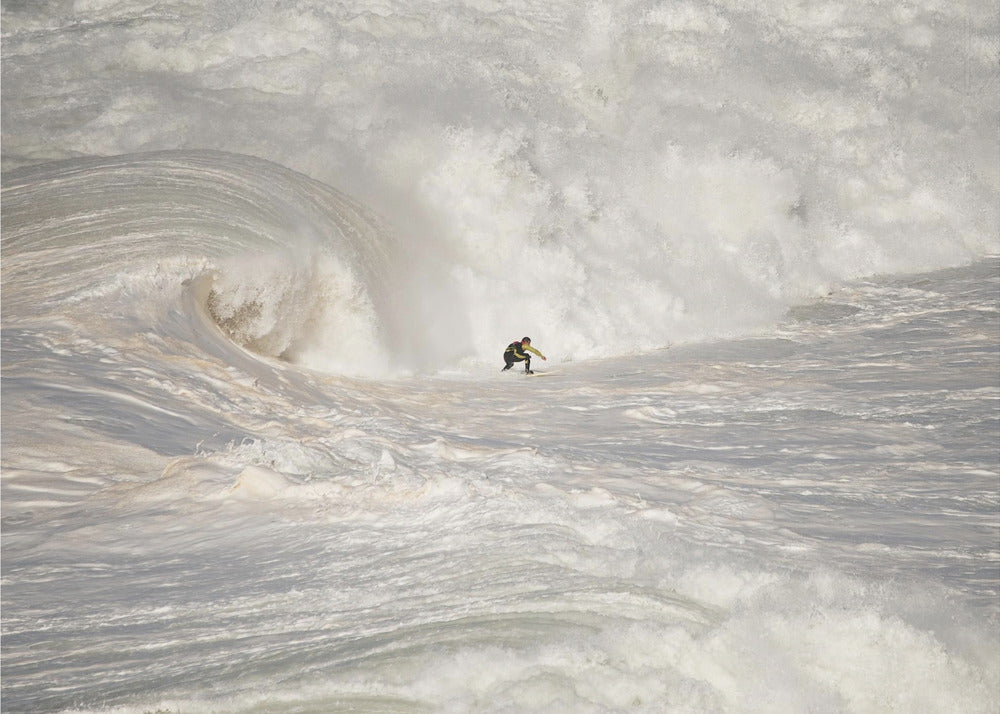 A framed photograph of a lone surfer dwarfed by a massive, powerful ocean wave. The water is a churning mass of white foam and spray, and the surfer is navigating the enormous swell. Print