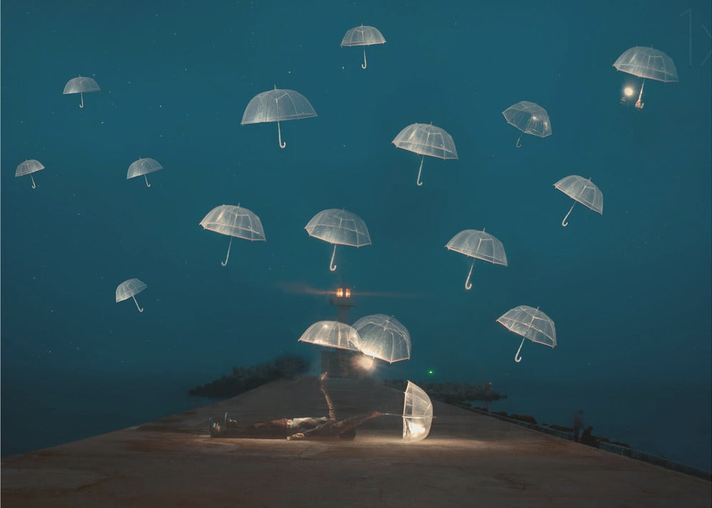 A surreal night scene with a person lying on a pier next to a glowing, transparent umbrella, while numerous other glowing umbrellas float like jellyfish in the dark blue sky above the sea, with a distant lighthouse in the background. Poster