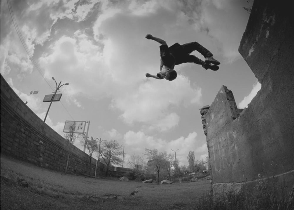A dynamic black and white fisheye photograph of a person mid-flip, jumping over a crumbling wall in an urban setting under a cloudy sky. The image has a silver-colored frame. Print