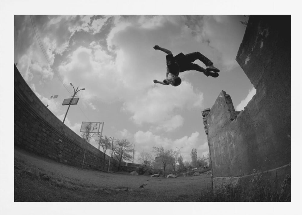 A dynamic black and white fisheye photograph of a person mid-flip, jumping over a crumbling wall in an urban setting under a cloudy sky. The image has a silver-colored frame. Print