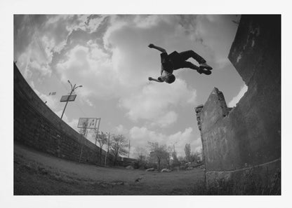 A dynamic black and white fisheye photograph of a person mid-flip, jumping over a crumbling wall in an urban setting under a cloudy sky. The image has a silver-colored frame. Print