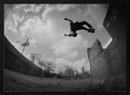 A dynamic black and white fisheye photograph of a person mid-flip, jumping over a crumbling wall in an urban setting under a cloudy sky. The image has a silver-colored frame. Print