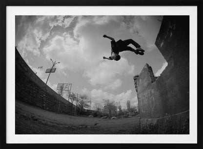 A dynamic black and white fisheye photograph of a person mid-flip, jumping over a crumbling wall in an urban setting under a cloudy sky. The image has a silver-colored frame. Print