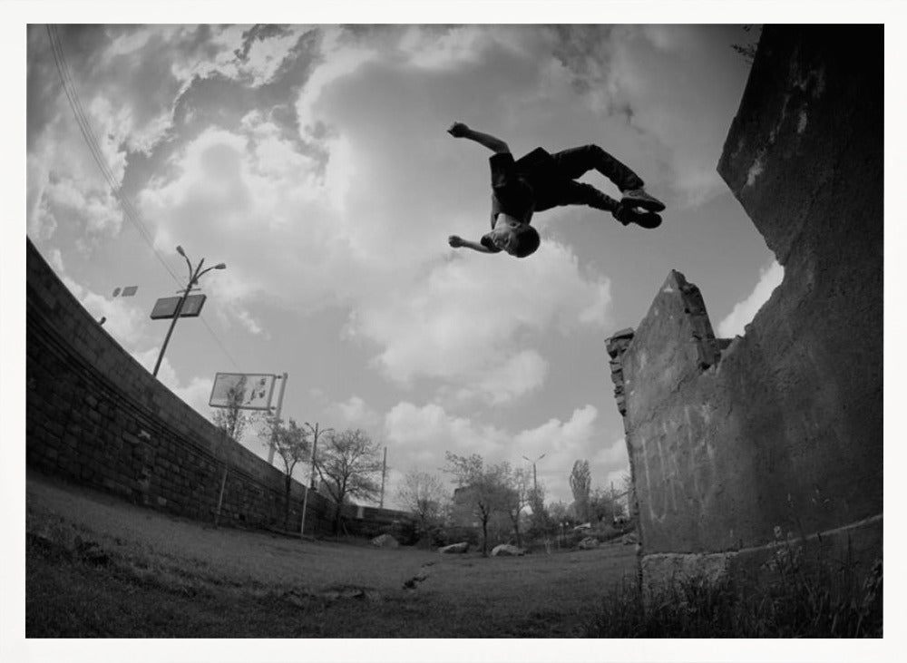 A dynamic black and white fisheye photograph of a person mid-flip, jumping over a crumbling wall in an urban setting under a cloudy sky. The image has a silver-colored frame. Print