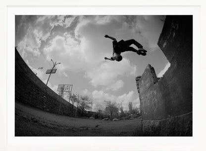 A dynamic black and white fisheye photograph of a person mid-flip, jumping over a crumbling wall in an urban setting under a cloudy sky. The image has a silver-colored frame. Print