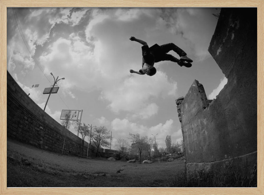 A dynamic black and white fisheye photograph of a person mid-flip, jumping over a crumbling wall in an urban setting under a cloudy sky. The image has a silver-colored frame. Print