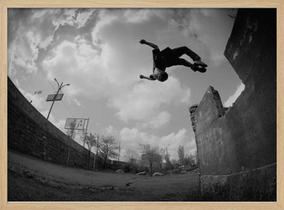 A dynamic black and white fisheye photograph of a person mid-flip, jumping over a crumbling wall in an urban setting under a cloudy sky. The image has a silver-colored frame. Print