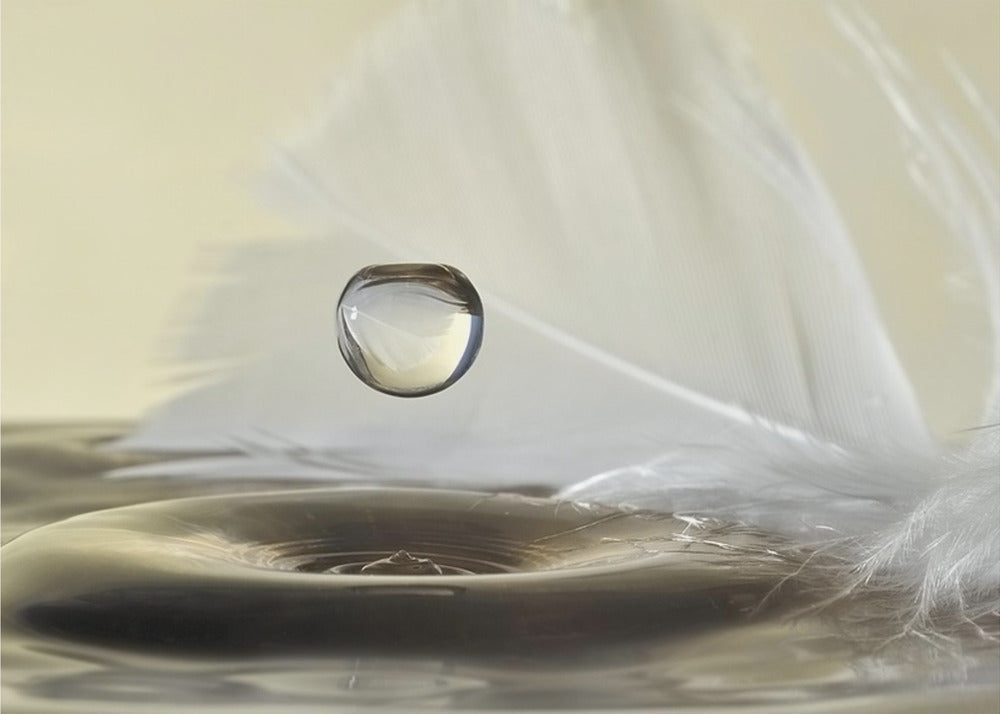 A high-speed macro photograph captures a perfectly spherical water droplet suspended in mid-air above a ripple on the water's surface. A soft white feather lies in the background, creating a serene and delicate composition with a neutral color palette, all enclosed in a brushed silver frame. Wall Art