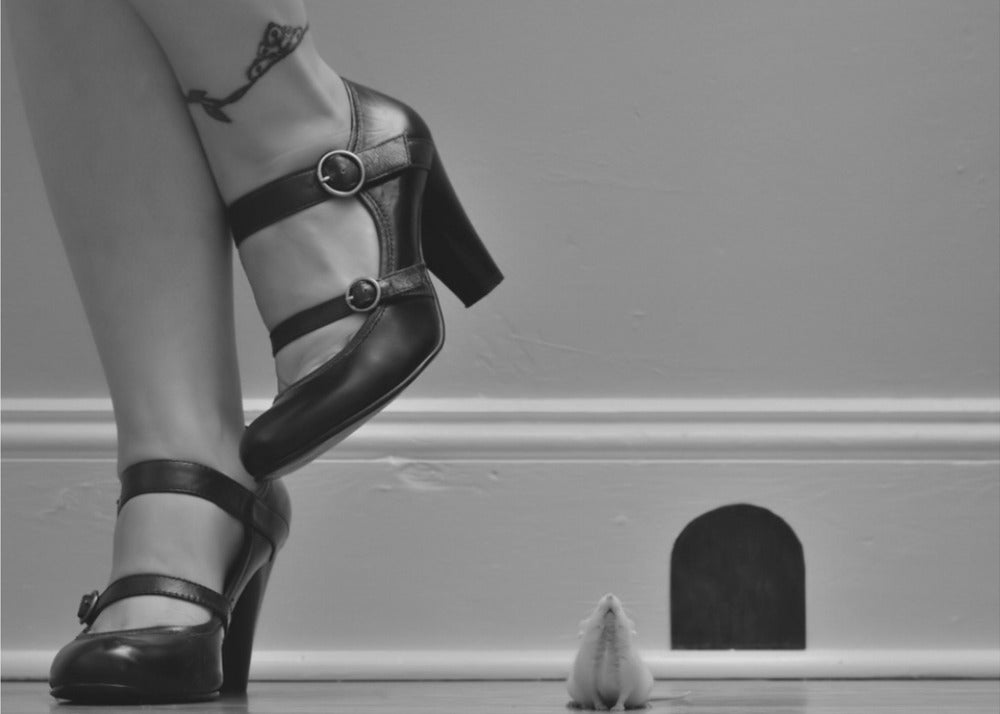 A black and white photograph showing the lower legs and feet of a woman wearing black high-heeled Mary Jane shoes. A tiny white mouse stands on its hind legs near a cartoonish mouse hole in the baseboard, looking up at the woman. Artwork