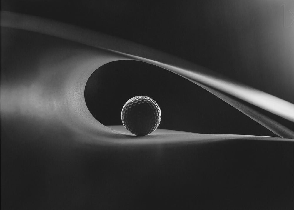 An artistic black and white photograph of a golf ball resting inside the gentle curve of a dark, abstract shape, creating a minimalist and dramatic composition with strong light and shadows. Poster