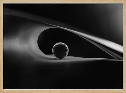 An artistic black and white photograph of a golf ball resting inside the gentle curve of a dark, abstract shape, creating a minimalist and dramatic composition with strong light and shadows. Poster