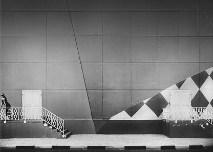A framed black and white photograph of a modern urban building. A woman in a dark outfit stands on the left by a white ornate railing and door. The large gray wall behind her is bisected by strong diagonal lines and features a bold black and white diamond pattern on the right, where another door and railing are visible. Decor