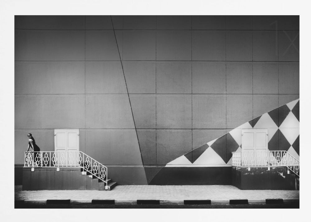 A framed black and white photograph of a modern urban building. A woman in a dark outfit stands on the left by a white ornate railing and door. The large gray wall behind her is bisected by strong diagonal lines and features a bold black and white diamond pattern on the right, where another door and railing are visible. Decor