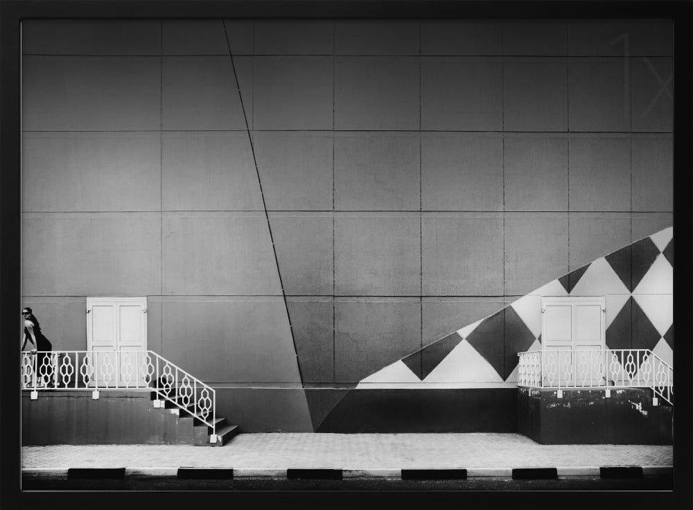 A framed black and white photograph of a modern urban building. A woman in a dark outfit stands on the left by a white ornate railing and door. The large gray wall behind her is bisected by strong diagonal lines and features a bold black and white diamond pattern on the right, where another door and railing are visible. Decor