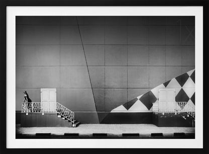 A framed black and white photograph of a modern urban building. A woman in a dark outfit stands on the left by a white ornate railing and door. The large gray wall behind her is bisected by strong diagonal lines and features a bold black and white diamond pattern on the right, where another door and railing are visible. Decor