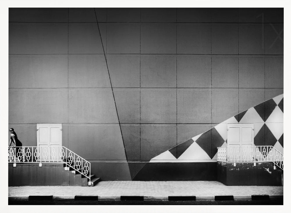 A framed black and white photograph of a modern urban building. A woman in a dark outfit stands on the left by a white ornate railing and door. The large gray wall behind her is bisected by strong diagonal lines and features a bold black and white diamond pattern on the right, where another door and railing are visible. Decor
