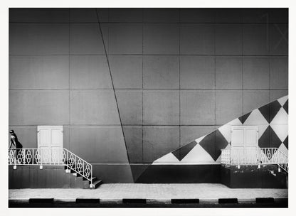 A framed black and white photograph of a modern urban building. A woman in a dark outfit stands on the left by a white ornate railing and door. The large gray wall behind her is bisected by strong diagonal lines and features a bold black and white diamond pattern on the right, where another door and railing are visible. Decor