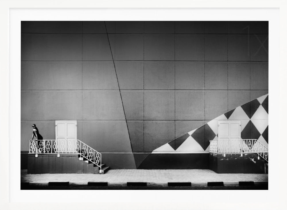 A framed black and white photograph of a modern urban building. A woman in a dark outfit stands on the left by a white ornate railing and door. The large gray wall behind her is bisected by strong diagonal lines and features a bold black and white diamond pattern on the right, where another door and railing are visible. Decor