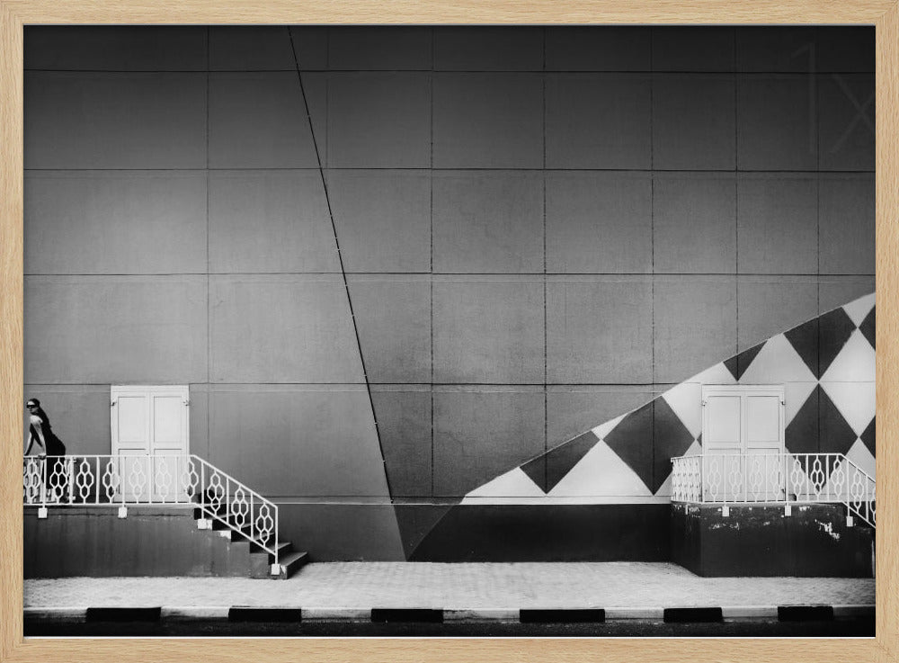 A framed black and white photograph of a modern urban building. A woman in a dark outfit stands on the left by a white ornate railing and door. The large gray wall behind her is bisected by strong diagonal lines and features a bold black and white diamond pattern on the right, where another door and railing are visible. Decor
