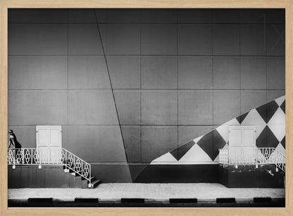 A framed black and white photograph of a modern urban building. A woman in a dark outfit stands on the left by a white ornate railing and door. The large gray wall behind her is bisected by strong diagonal lines and features a bold black and white diamond pattern on the right, where another door and railing are visible. Decor