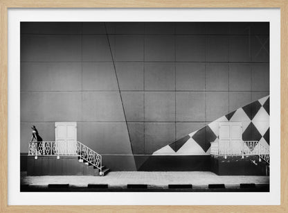 A framed black and white photograph of a modern urban building. A woman in a dark outfit stands on the left by a white ornate railing and door. The large gray wall behind her is bisected by strong diagonal lines and features a bold black and white diamond pattern on the right, where another door and railing are visible. Decor