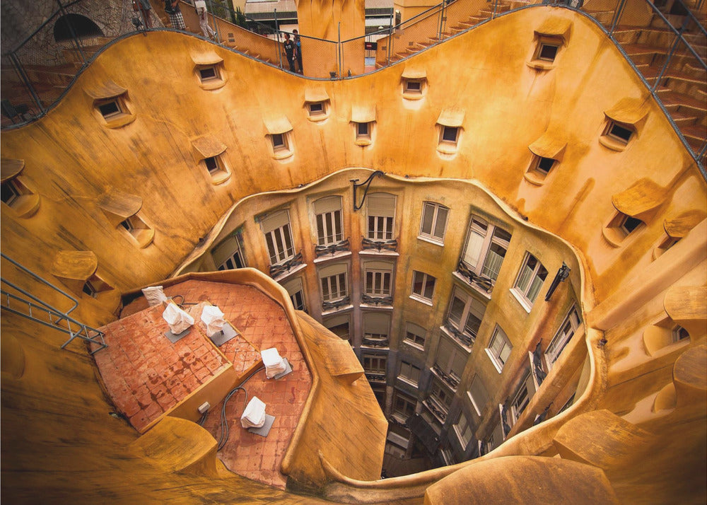 A high-angle photograph looking down into the surreal, undulating inner courtyard of an architectural masterpiece. The warm, golden-ochre walls curve organically, dotted with small, hooded windows, encircling the deep central lightwell that reveals floors of apartments below. Wall Art
