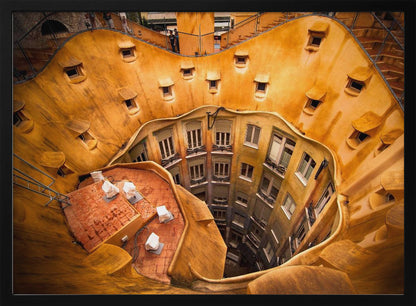 A high-angle photograph looking down into the surreal, undulating inner courtyard of an architectural masterpiece. The warm, golden-ochre walls curve organically, dotted with small, hooded windows, encircling the deep central lightwell that reveals floors of apartments below. Wall Art