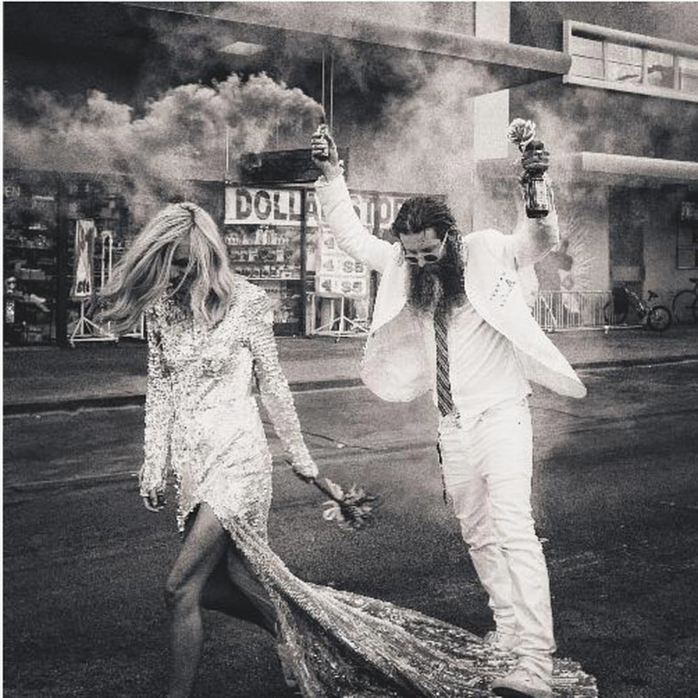 A black and white photograph in a light wood frame showing a stylish couple walking down a street. The woman wears a sparkling sequin gown, and the man, in a white suit with a long beard, triumphantly raises his arms, holding a smoke bomb and a bottle with a rose. Decor