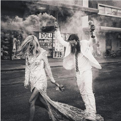 A black and white photograph in a light wood frame showing a stylish couple walking down a street. The woman wears a sparkling sequin gown, and the man, in a white suit with a long beard, triumphantly raises his arms, holding a smoke bomb and a bottle with a rose. Decor