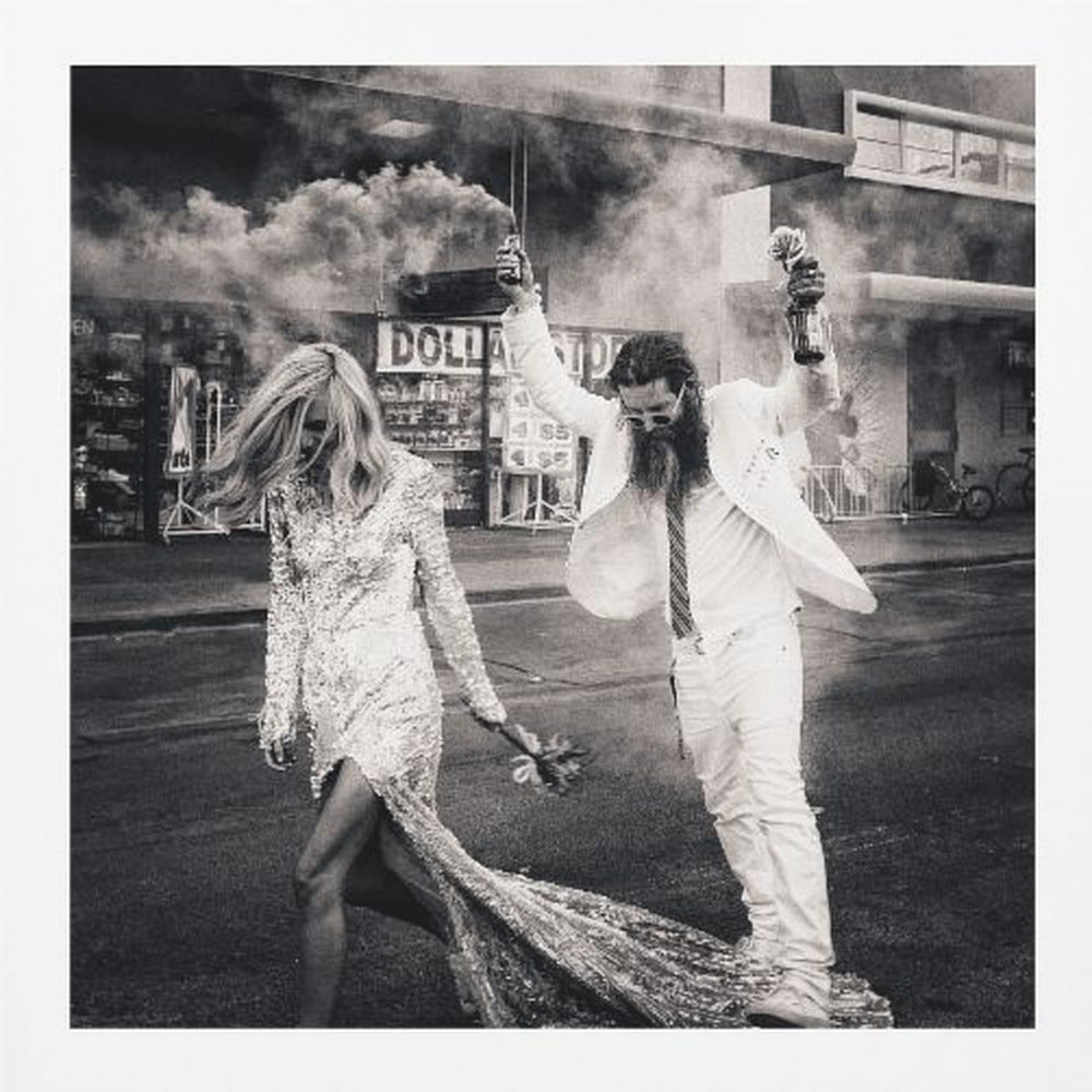 A black and white photograph in a light wood frame showing a stylish couple walking down a street. The woman wears a sparkling sequin gown, and the man, in a white suit with a long beard, triumphantly raises his arms, holding a smoke bomb and a bottle with a rose. Decor