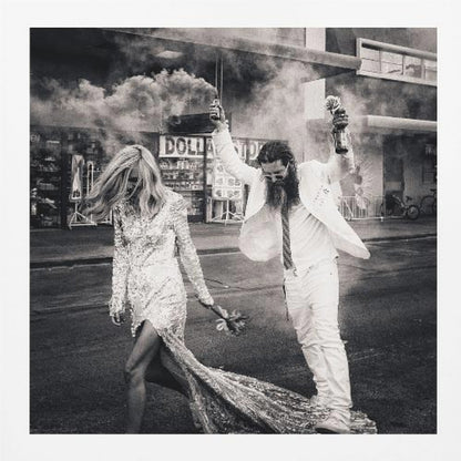 A black and white photograph in a light wood frame showing a stylish couple walking down a street. The woman wears a sparkling sequin gown, and the man, in a white suit with a long beard, triumphantly raises his arms, holding a smoke bomb and a bottle with a rose. Decor