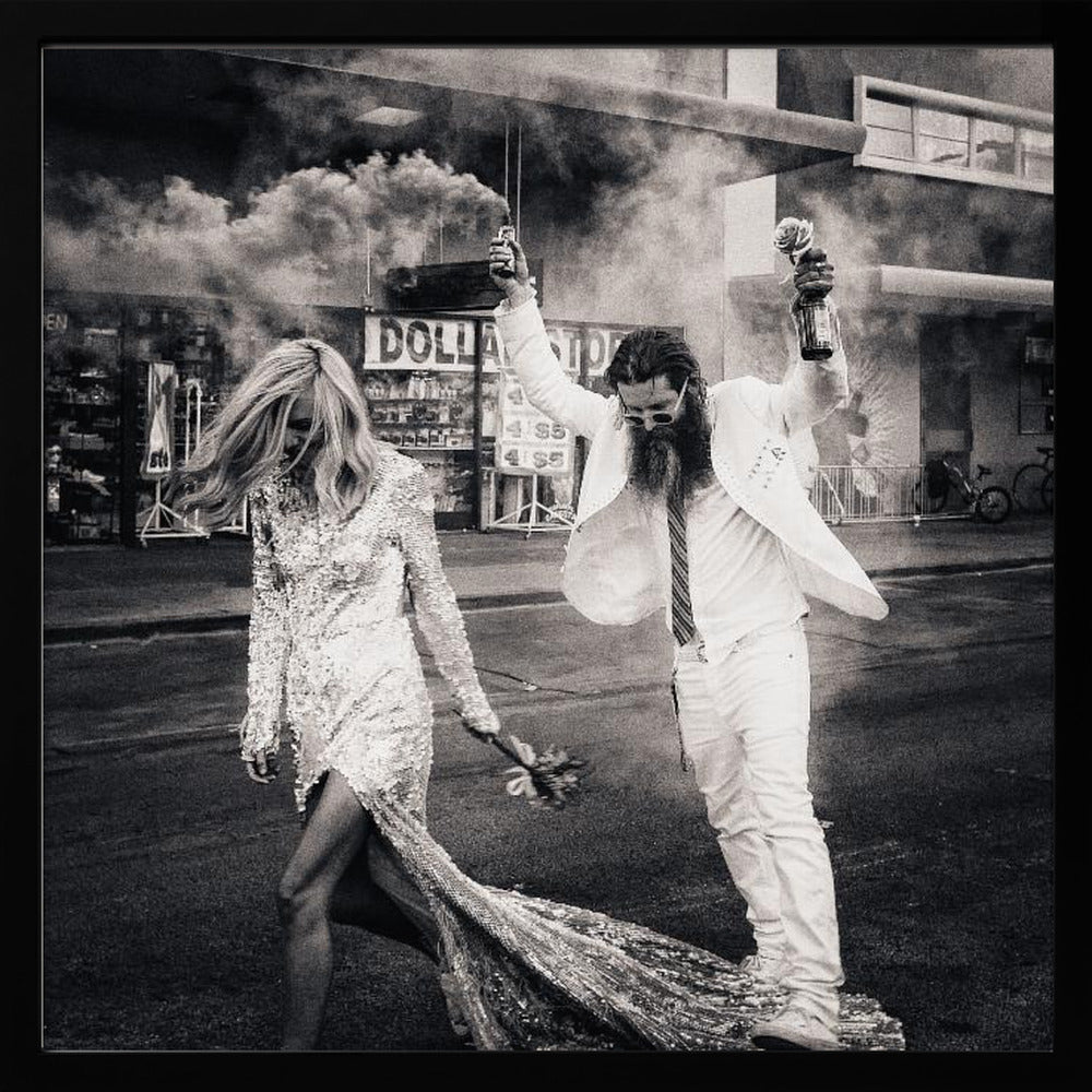 A black and white photograph in a light wood frame showing a stylish couple walking down a street. The woman wears a sparkling sequin gown, and the man, in a white suit with a long beard, triumphantly raises his arms, holding a smoke bomb and a bottle with a rose. Decor