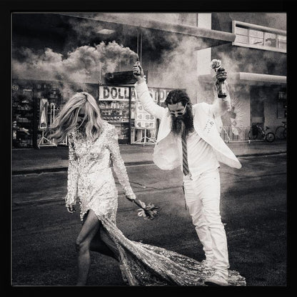 A black and white photograph in a light wood frame showing a stylish couple walking down a street. The woman wears a sparkling sequin gown, and the man, in a white suit with a long beard, triumphantly raises his arms, holding a smoke bomb and a bottle with a rose. Decor