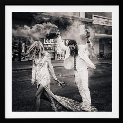 A black and white photograph in a light wood frame showing a stylish couple walking down a street. The woman wears a sparkling sequin gown, and the man, in a white suit with a long beard, triumphantly raises his arms, holding a smoke bomb and a bottle with a rose. Decor