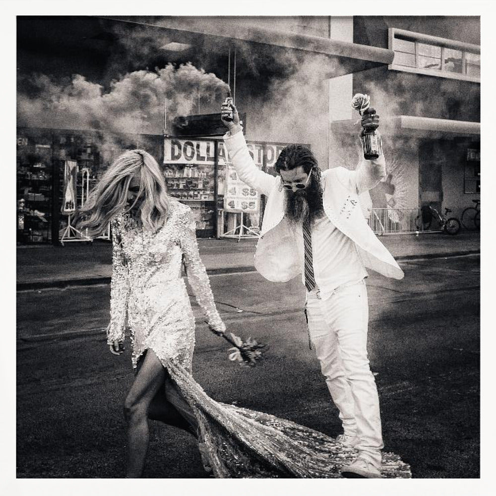 A black and white photograph in a light wood frame showing a stylish couple walking down a street. The woman wears a sparkling sequin gown, and the man, in a white suit with a long beard, triumphantly raises his arms, holding a smoke bomb and a bottle with a rose. Decor