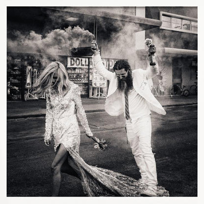 A black and white photograph in a light wood frame showing a stylish couple walking down a street. The woman wears a sparkling sequin gown, and the man, in a white suit with a long beard, triumphantly raises his arms, holding a smoke bomb and a bottle with a rose. Decor